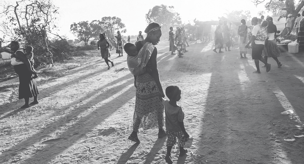 A woman walks among a crowd, holding the hand of a toddler and carrying a baby.