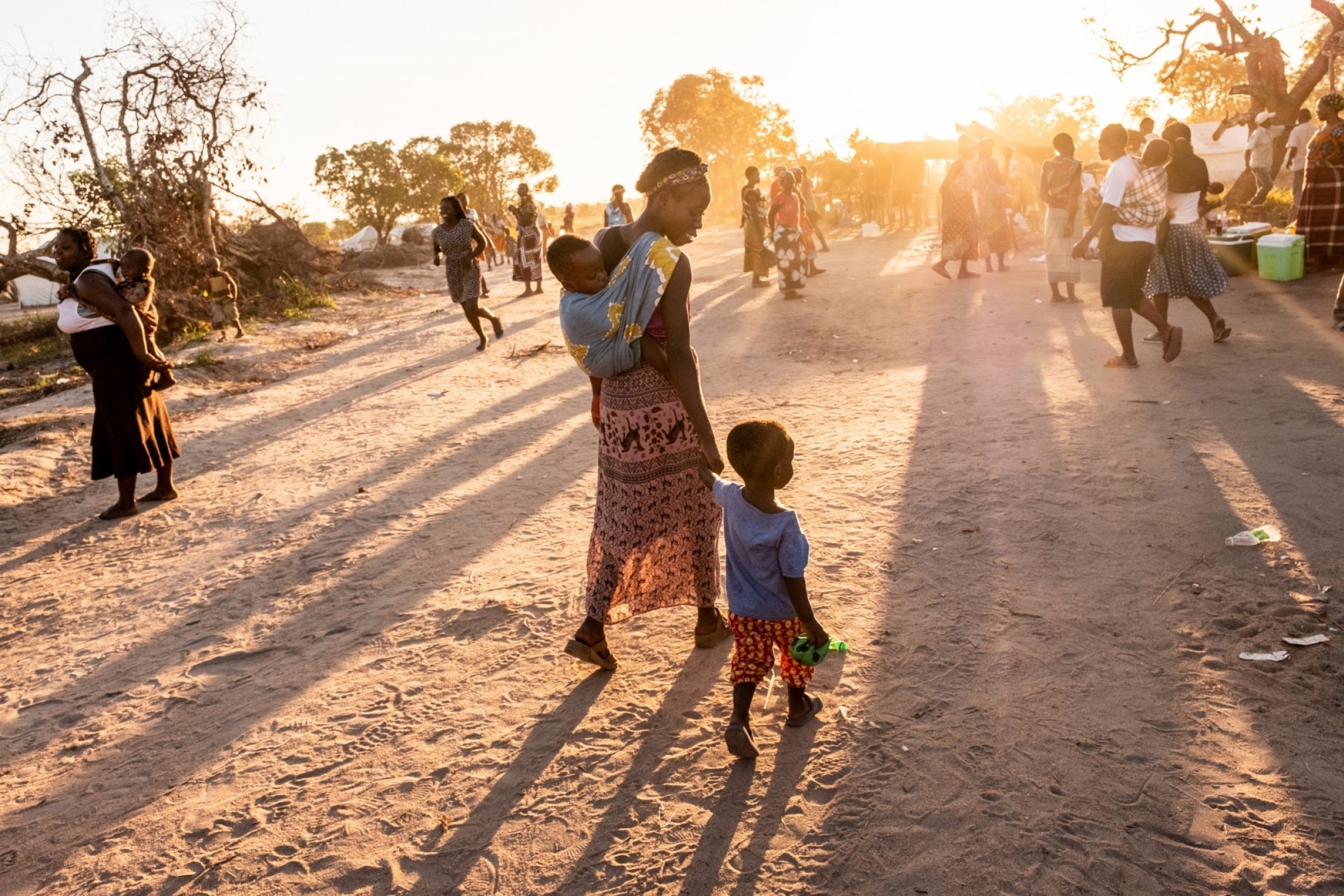 A woman walks among a crowd, holding the hand of a toddler and carrying a baby.