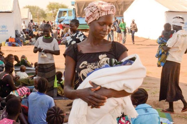 Person in a headwrap stands outside in a crowd looking down at their child.